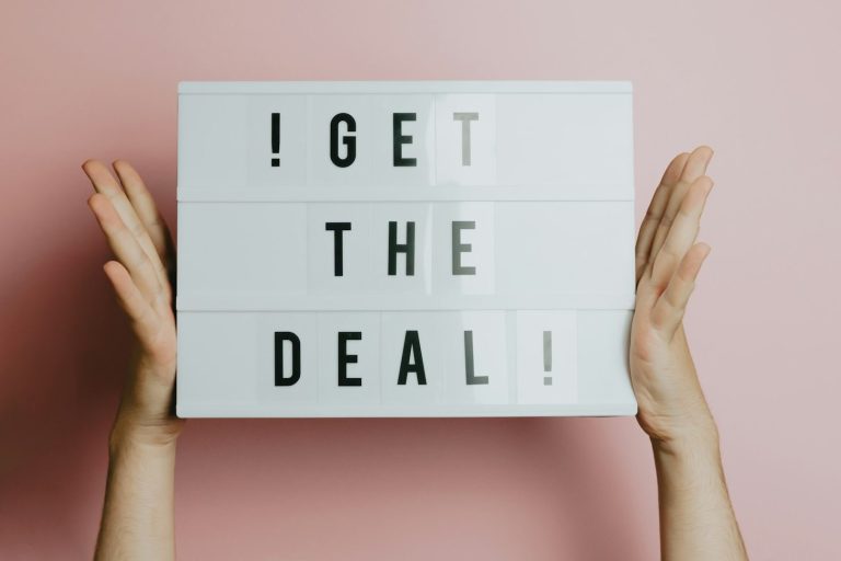 Get the Deal Bild zur Seite Verkauf Hände halten ein Schild mit der Aufschrift „GET THE DEAL!“ vor einem rosa Hintergrund.