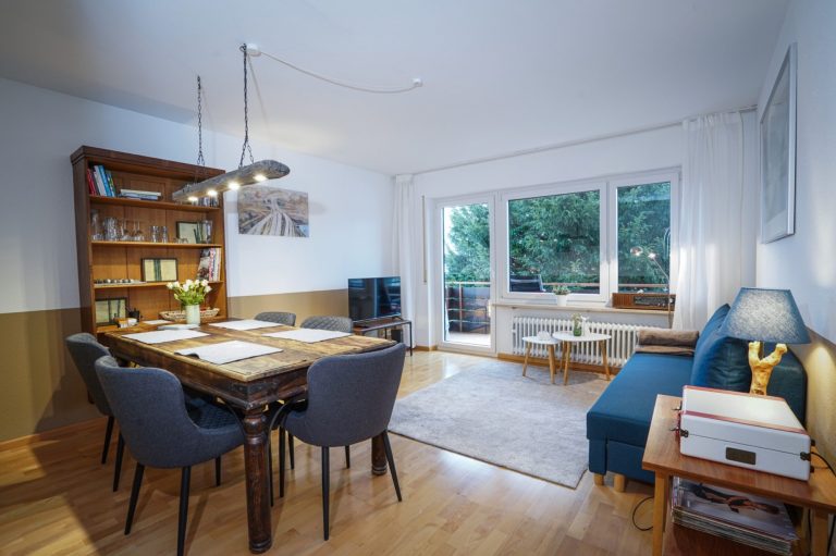Wohnzimmer zur Seite Miete Helles Wohnzimmer mit Esstisch, Stühlen, Bücherregal und einer Couch. Großer Fensterbereich.