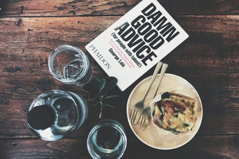 Passendes Buch zur Seite Beratung Ein Buch mit dem Titel "Damn Good Advice" liegt neben einem Teller und Gläsern auf einem Holztisch.